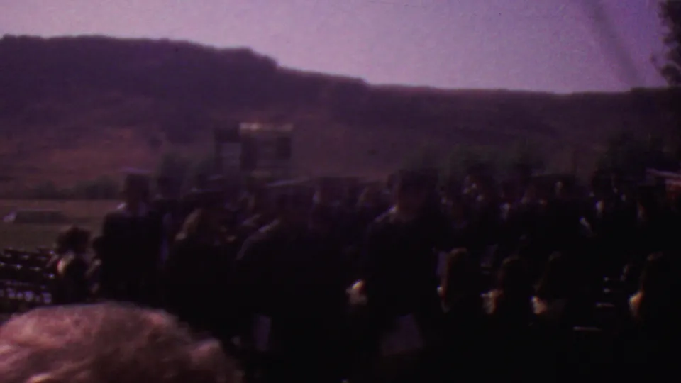 Wide 8mm view of the outdoor graduation ceremony, with attendees and graduates in shadow, rows of seating, and low hills beyond the grounds.
