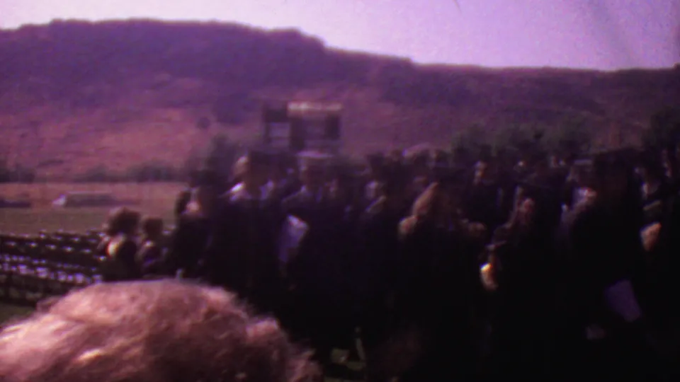 Outdoor graduation crowd seen from a spectator position, with dark-robed figures, a small ceremony structure, foreground heads, and hills behind the event area.