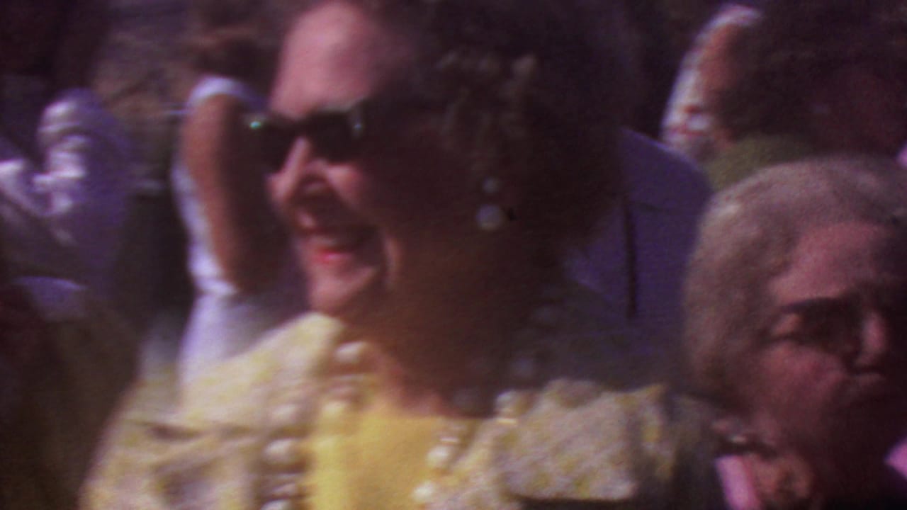 1965 Los Angeles home-movie frame showing adults at an outdoor gathering, with a woman in sunglasses and large pearls near a man in a dark suit.