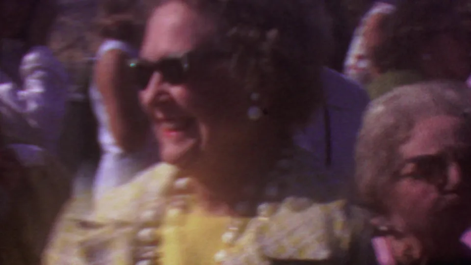 1965 Los Angeles home-movie frame showing adults at an outdoor gathering, with a woman in sunglasses and large pearls near a man in a dark suit.