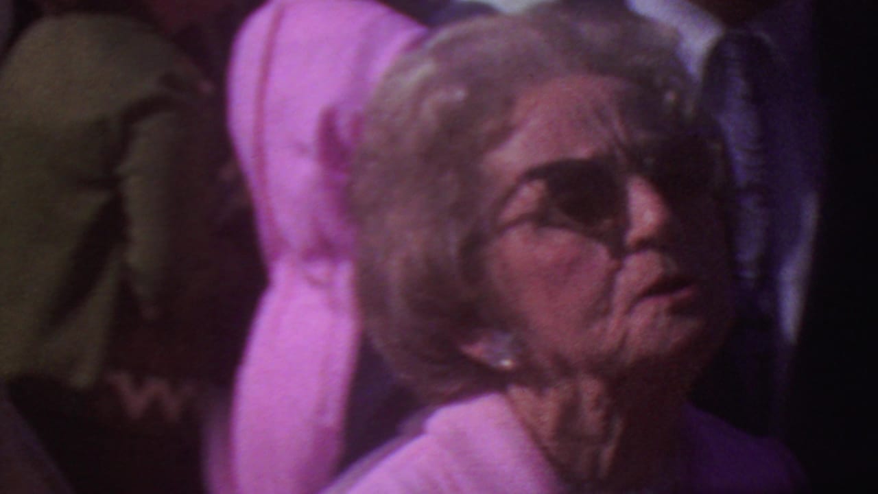 Older women sit or stand close together in a crowd, including one in a pink outfit and sunglasses.