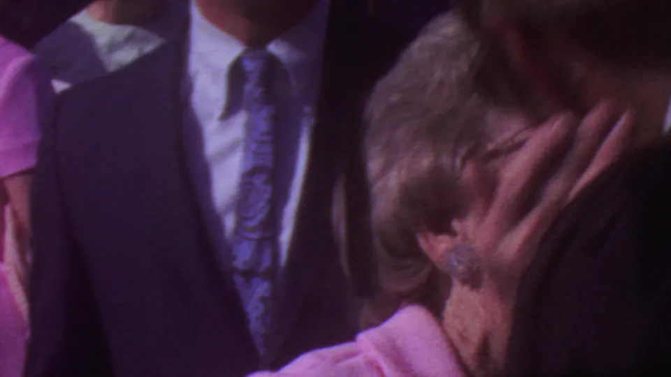 1965 Los Angeles home-movie frame showing a suited man and a woman in pink, with the woman lifting a hand near her face amid a crowded outdoor group.