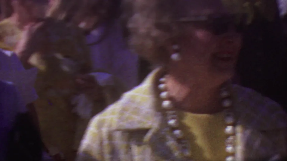1965 Los Angeles home-movie frame showing a woman in sunglasses, a light patterned coat, yellow top, and large pearls among adults at an outdoor event.