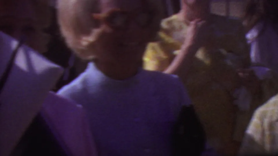 Close handheld 1965 Los Angeles graduation crowd view with adults in period clothing and papers partly filling the foreground.