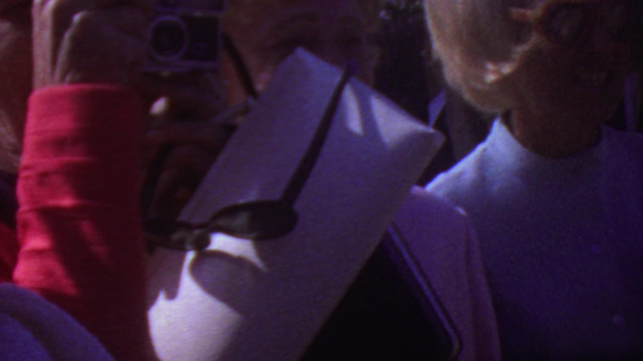 Handheld graduation crowd shot in Los Angeles, 1965, with a person raising a small camera near paper programs and nearby attendees.