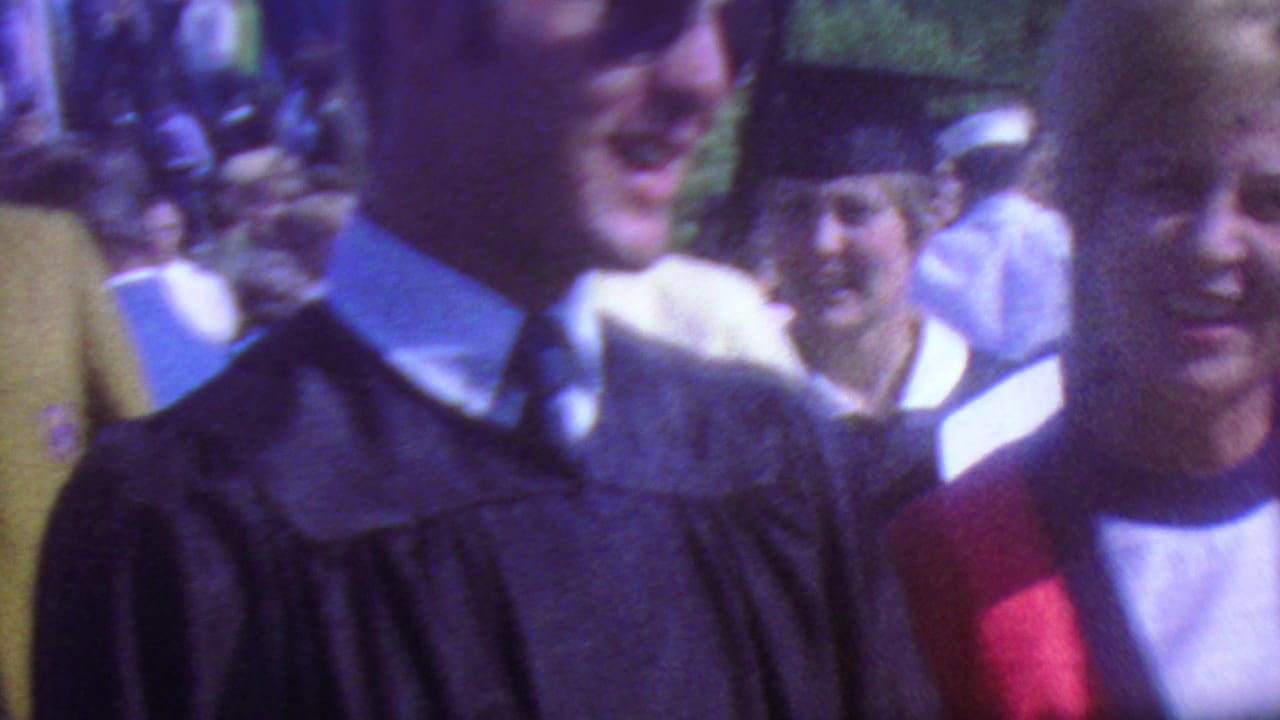 Graduate in dark gown stands near a woman in a red stole and other smiling attendees.
