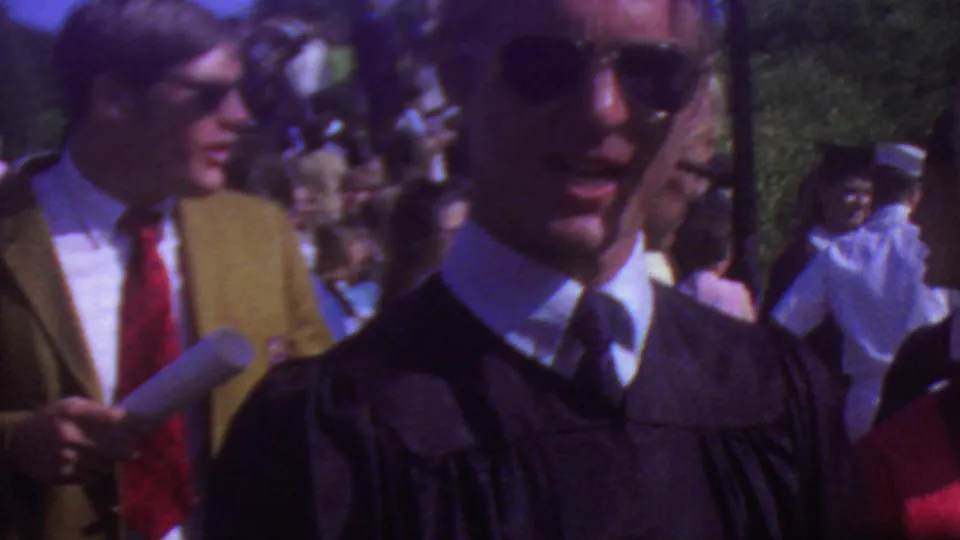 Outdoor 1965 Los Angeles graduation crowd with a man in a black gown and sunglasses near attendees, papers, and a handheld microphone.