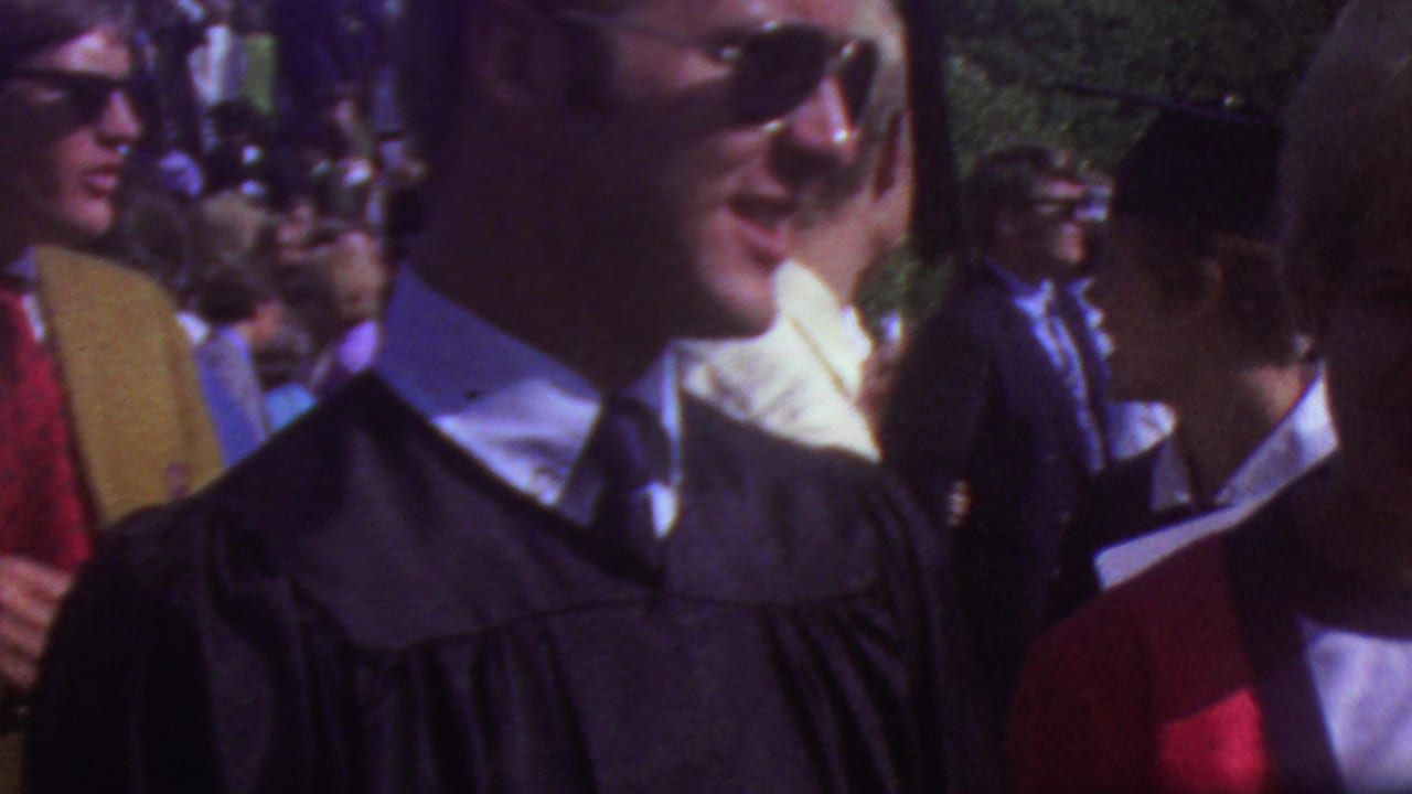 Young men in black graduation caps and gowns stand close to visitors in an outdoor Los Angeles graduation crowd from the 1965 home-movie footage.