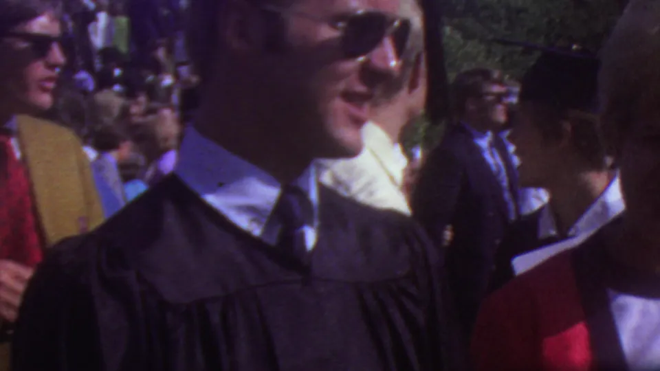 Young men in black graduation caps and gowns stand close to visitors in an outdoor Los Angeles graduation crowd from the 1965 home-movie footage.