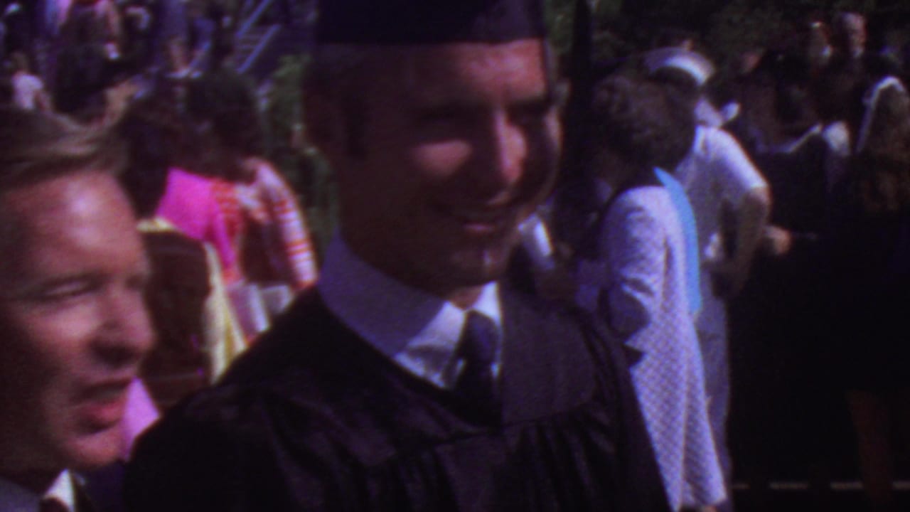 A smiling graduate in black academic regalia appears beside a man in a red jacket as people mingle outdoors in the 1965 Los Angeles graduation footage.