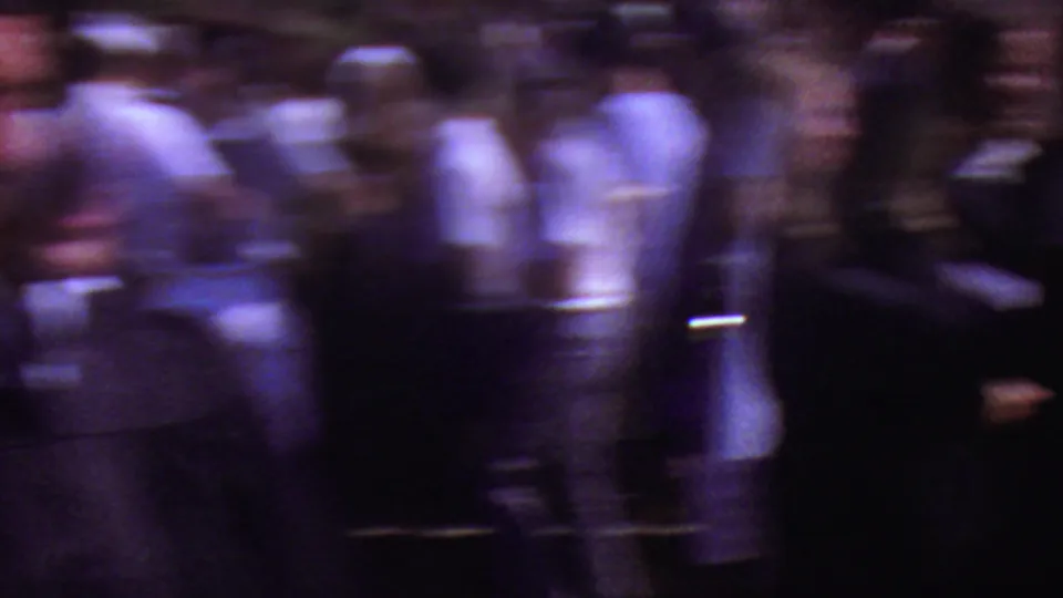 A figure in dark graduation regalia moves past visitors in a soft, blurred 1965 Los Angeles outdoor commencement crowd.