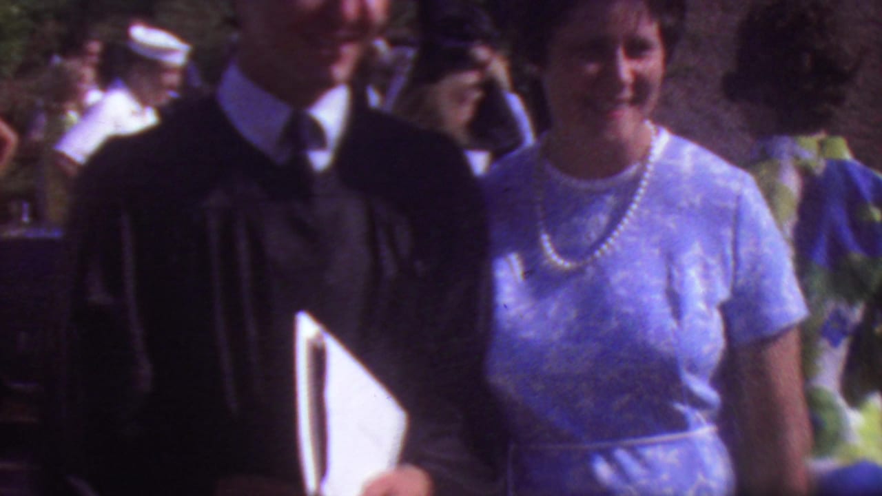 In 1965 Los Angeles home-movie footage, a graduate in a black academic gown stands beside a woman in a light blue dress and pearl necklace as guests gather outdoors.