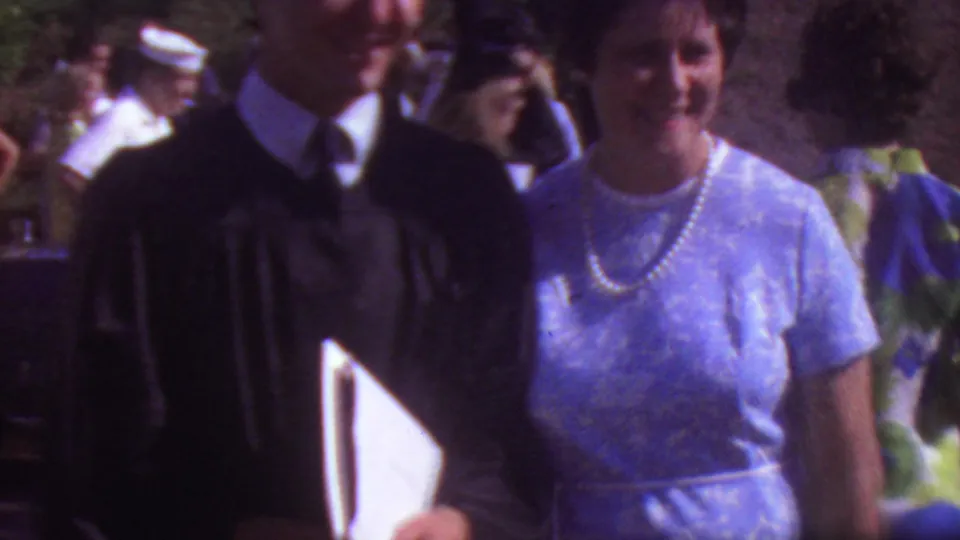 In 1965 Los Angeles home-movie footage, a graduate in a black academic gown stands beside a woman in a light blue dress and pearl necklace as guests gather outdoors.