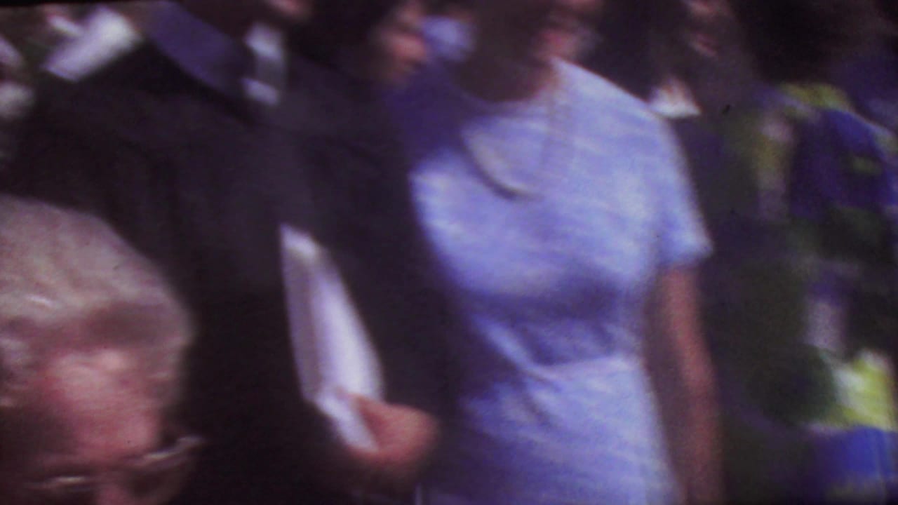 Blurred close view of a woman in a light blue dress walking through a graduation crowd.