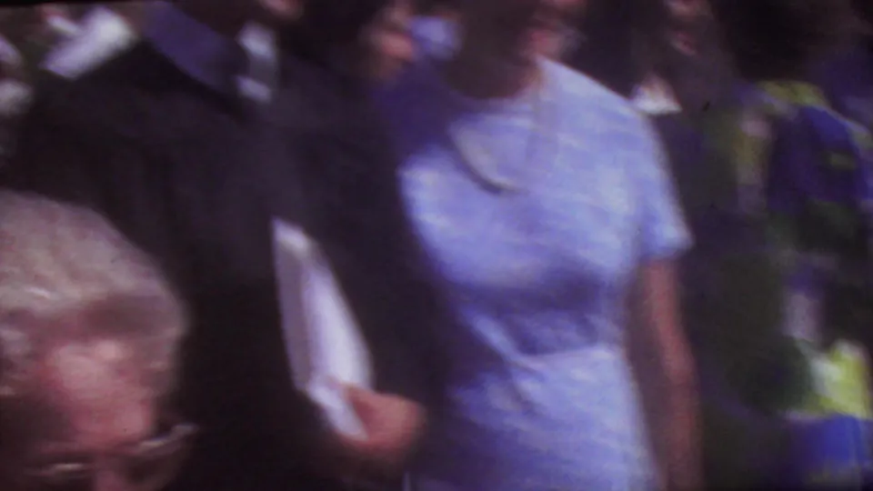 A woman in a light blue dress appears in motion within an outdoor graduation crowd, with academic caps and guests visible around her.