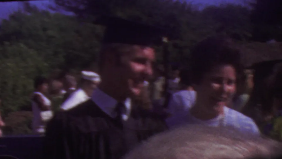 The graduate in cap and gown stands near a woman in a light blue dress, with another person passing close to the camera and guests visible behind them outdoors.