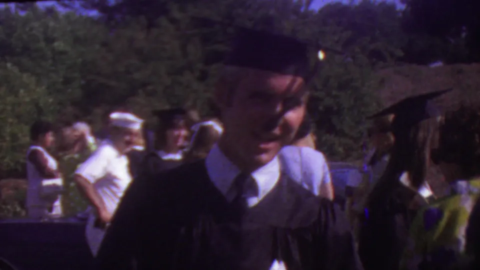 A smiling graduate in black academic cap and gown stands in the foreground as other graduates, guests, trees, and sunlit outdoor space appear behind him.