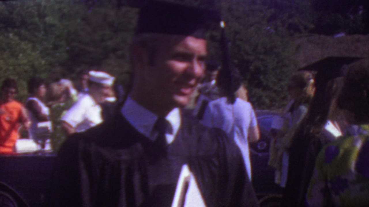 Young graduate in a black mortarboard and gown smiles toward the camera during an outdoor 1965 Los Angeles graduation gathering, with other people and cars behind him.