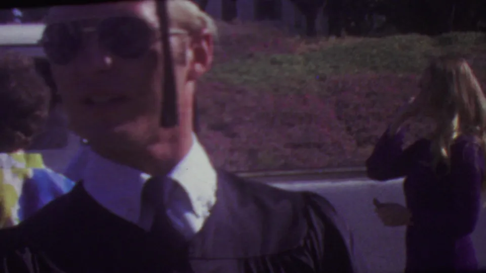 A young man in graduation regalia stands close to the lens at an outdoor Los Angeles graduation scene, with other cap-and-gown figures and guests visible in the background.