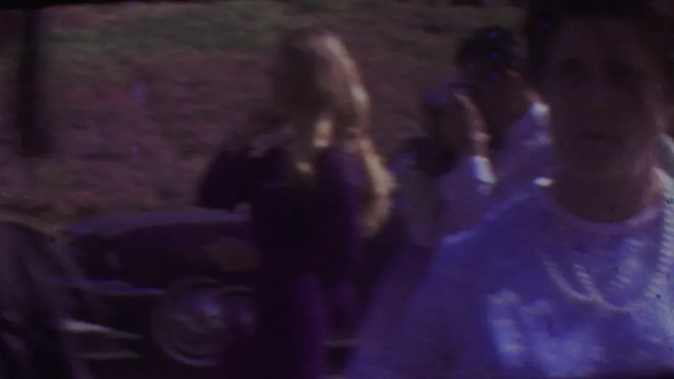 Two women in light-colored dresses with pearl necklaces pass close to the camera at the 1965 Los Angeles graduation gathering, with cars and guests behind them.