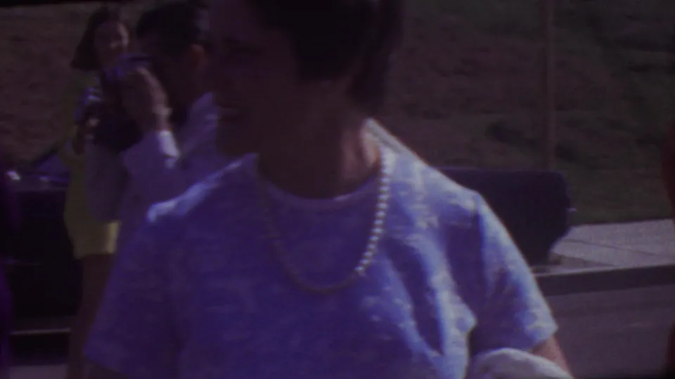 A woman in a light dress and pearl necklace is seen near a parked car at the outdoor Los Angeles graduation gathering, with other guests and a hillside behind her.
