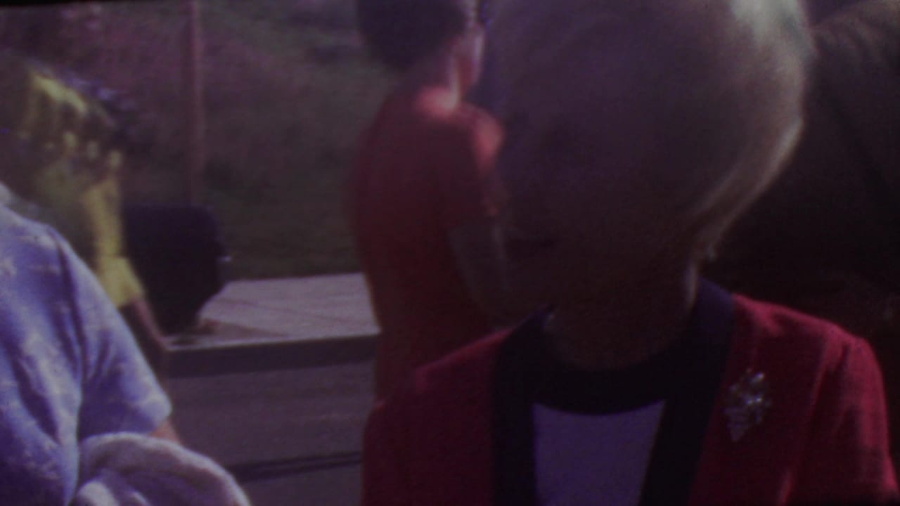 Soft 8mm frame from the 1965 Los Angeles graduation clip showing a woman in a red jacket near another adult in dressy clothing, with pavement and a hillside in the background.