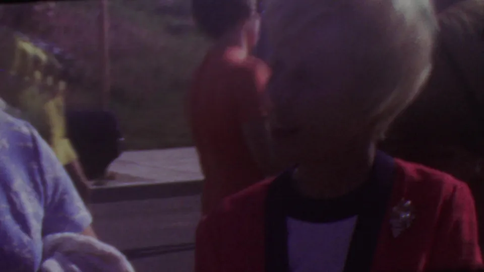 Soft 8mm frame from the 1965 Los Angeles graduation clip showing a woman in a red jacket near another adult in dressy clothing, with pavement and a hillside in the background.