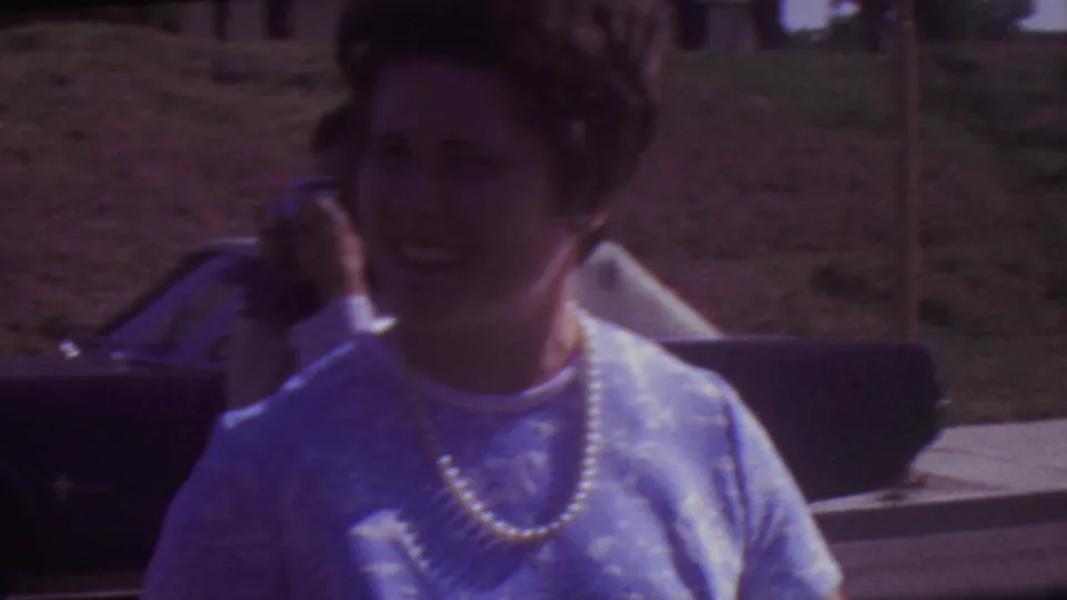 1965 Los Angeles graduation home-movie frame showing a woman in a blue patterned dress with a pearl necklace smiling outdoors, with cars and a hillside behind her.