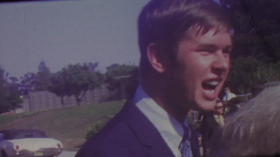 1965 Los Angeles graduation frame showing a young man in a dark suit and tie speaking or smiling beside another person, with trees, lawn, and a parked car in the background.