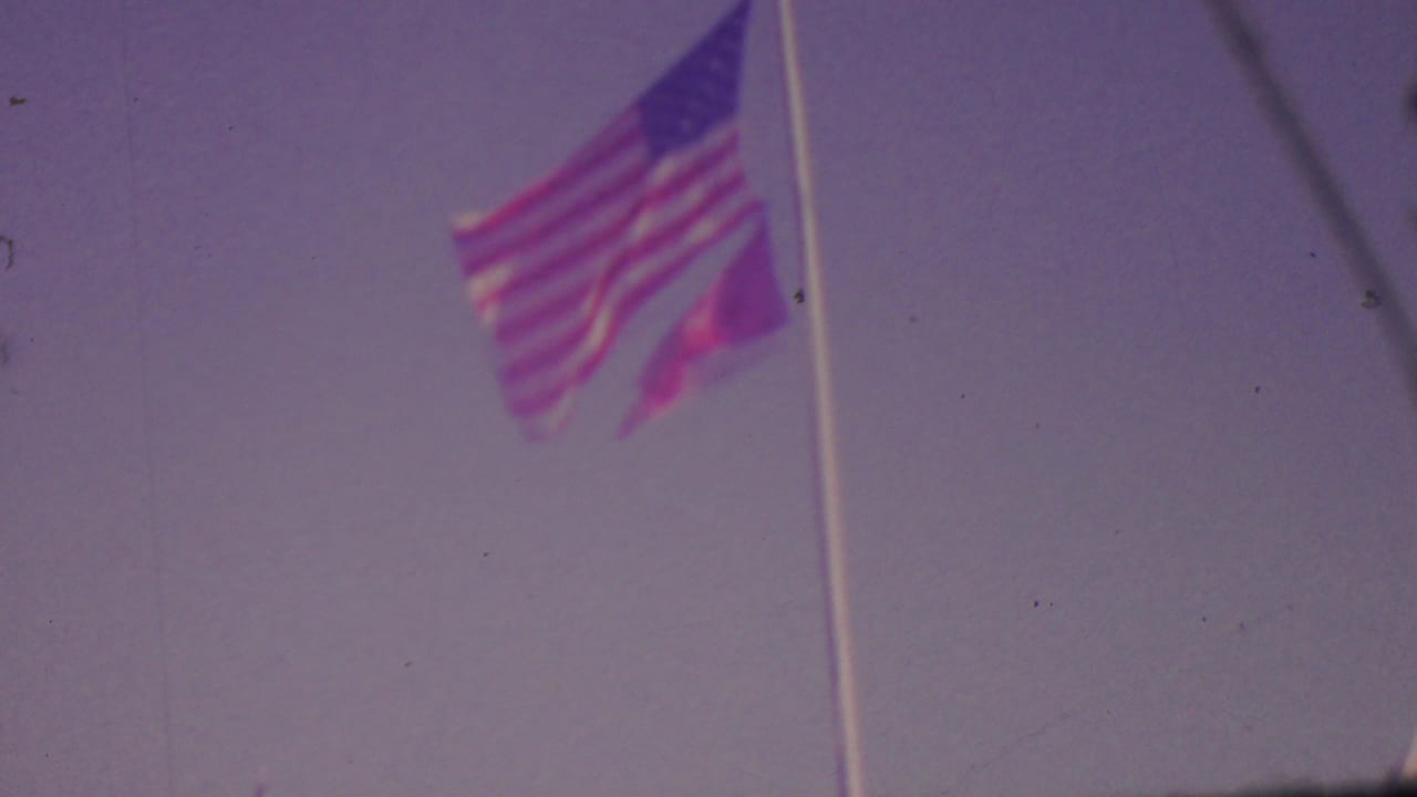 A U.S. flag waves on a tall pole against a pale sky in faded 8mm footage associated with the 1965 Los Angeles home-movie clip.