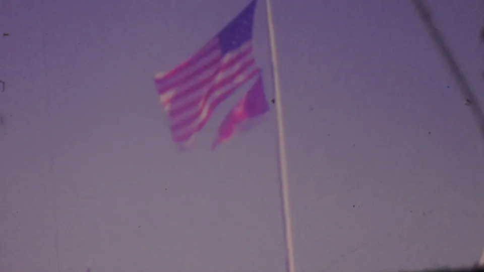 A U.S. flag waves on a tall pole against a pale sky in faded 8mm footage associated with the 1965 Los Angeles home-movie clip.