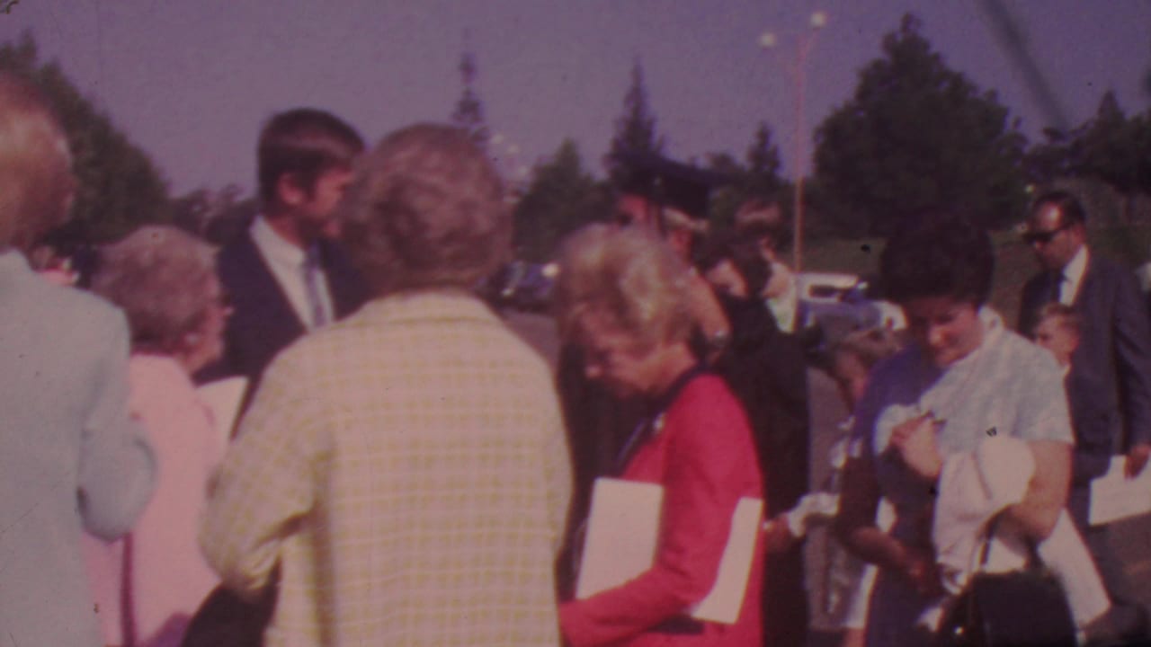 Adults gathered outdoors near parked cars, with a young woman in red holding papers.
