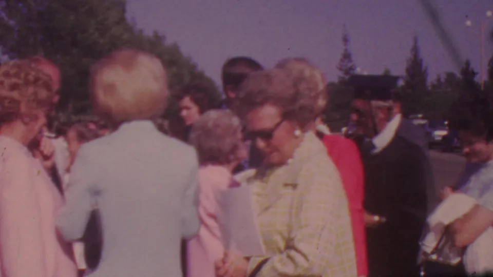 A handheld 8mm view shows adults in period clothing gathered outdoors near parked cars and trees during the Los Angeles celebration.