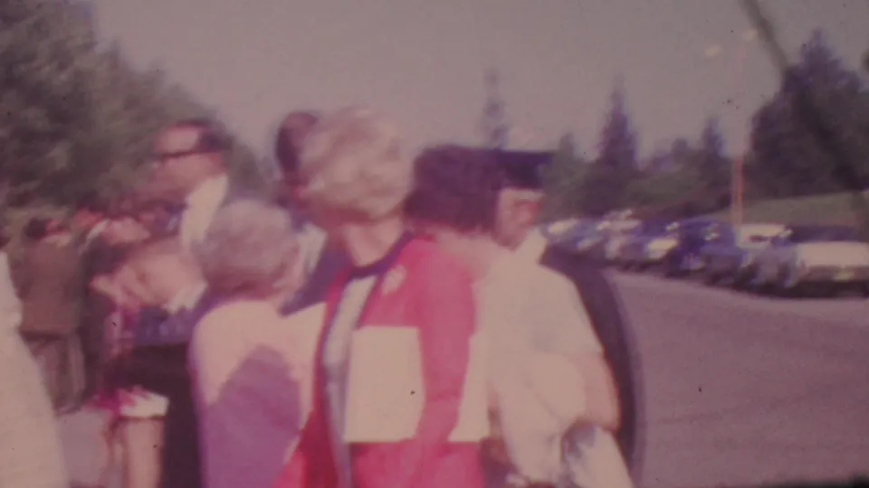 A young woman in a red cap and jacket stands among adults near a parking area, suggesting a graduation moment in the 1965 Los Angeles home movie.