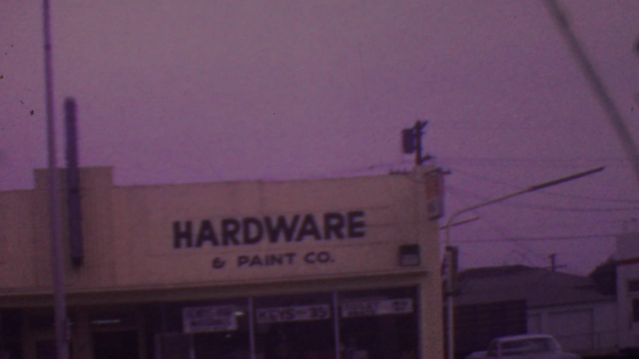 1965 Los Angeles street-level view of a white hardware and paint storefront with black lettering, glass windows, utility wires, and a purple-tinted 8mm look.
