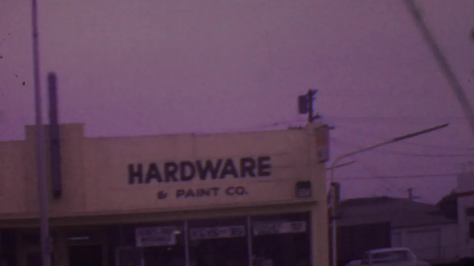 1965 Los Angeles street-level view of a white hardware and paint storefront with black lettering, glass windows, utility wires, and a purple-tinted 8mm look.