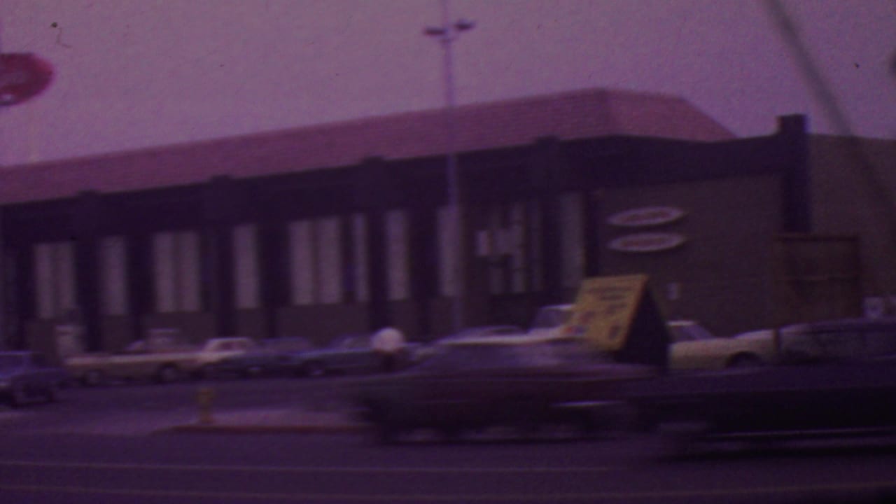 1965 Los Angeles roadside restaurant exterior seen from across the street, with parked mid-century cars, a red oval sign, and soft 8mm color shift.