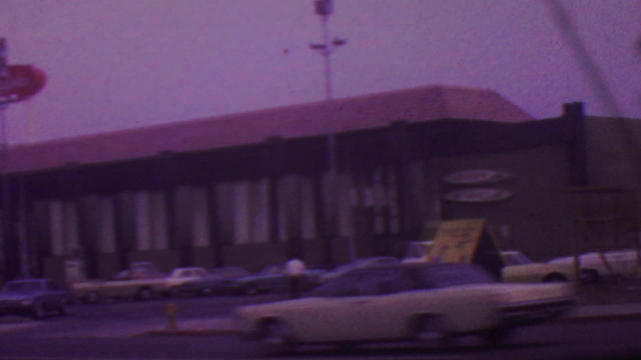 1965 Los Angeles street view of a low restaurant building with tall signage, parked 1960s cars, and a light-colored car moving through the foreground.