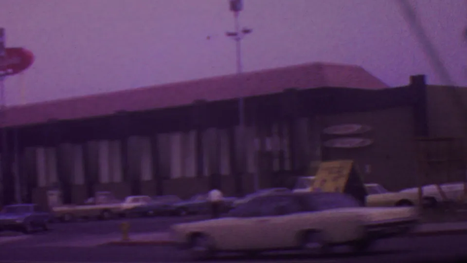 1965 Los Angeles street view of a low restaurant building with tall signage, parked 1960s cars, and a light-colored car moving through the foreground.
