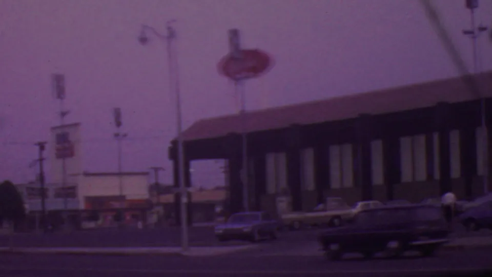 Wider 1965 Los Angeles home-movie view showing the roadside restaurant, pole signs, parked cars, moving traffic, and a pedestrian near the sidewalk.