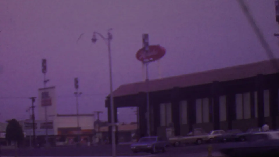 1965 Los Angeles roadside restaurant exterior with red oval signage above the roofline, several parked cars, and a blurred vehicle at the lower edge of frame.