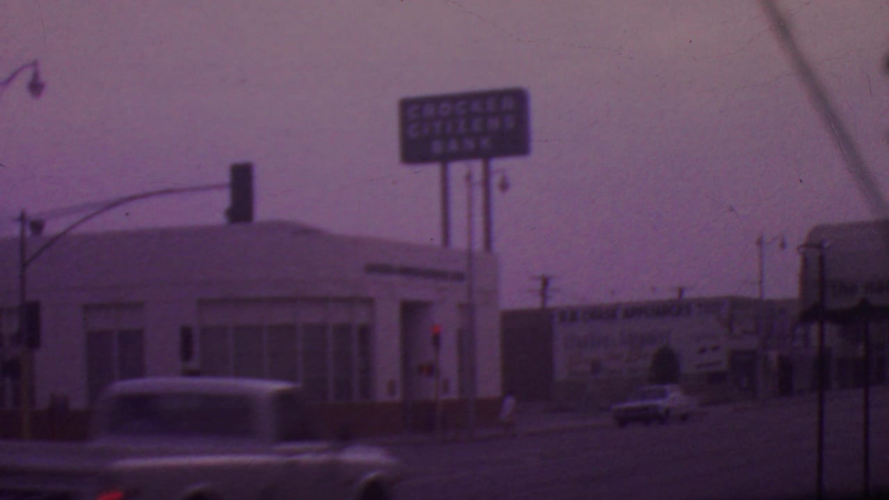 A blurred classic car crosses the foreground of a 1965 Los Angeles intersection with bank signage, traffic signals, and commercial billboards behind it.