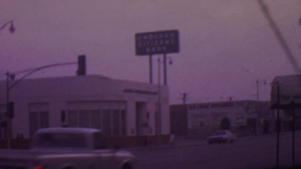 A blurred classic car crosses the foreground of a 1965 Los Angeles intersection with bank signage, traffic signals, and commercial billboards behind it.