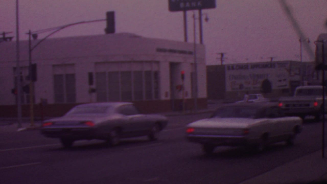 1965 Los Angeles street view with several cars at or near an intersection, overhead signals, commercial buildings, and distant business signage.