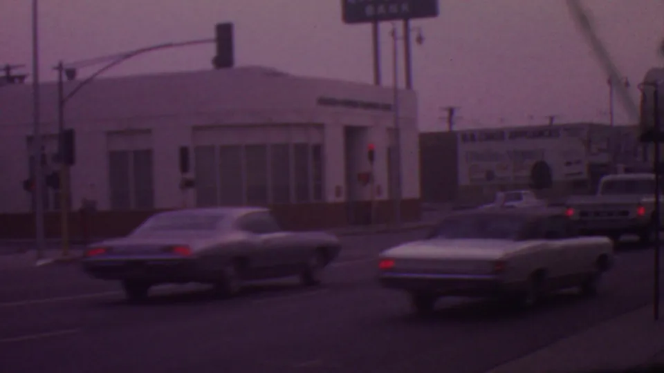 1965 Los Angeles street view with several cars at or near an intersection, overhead signals, commercial buildings, and distant business signage.