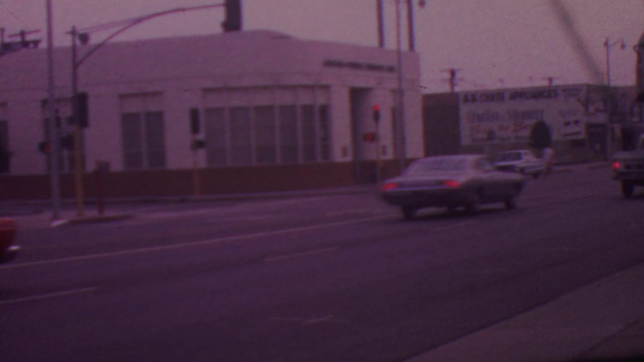 1965 Los Angeles intersection with traffic lights, a pedestrian crossing, several cars, and visible commercial signage including a bank and appliance billboard.