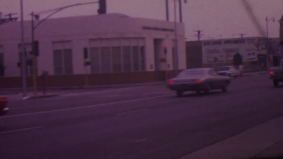 1965 Los Angeles intersection with traffic lights, a pedestrian crossing, several cars, and visible commercial signage including a bank and appliance billboard.