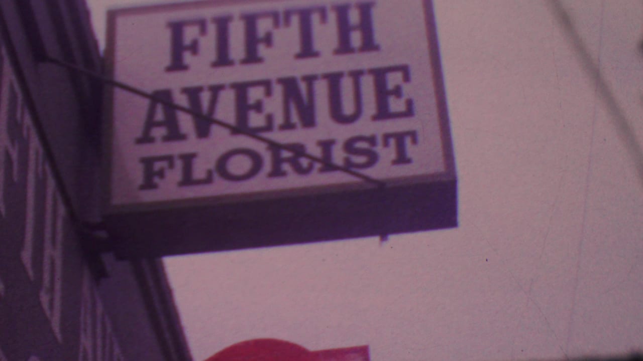 Close 8mm view of a Fifth Avenue Florist sign mounted above a sidewalk, with the building edge and tinted sky visible.