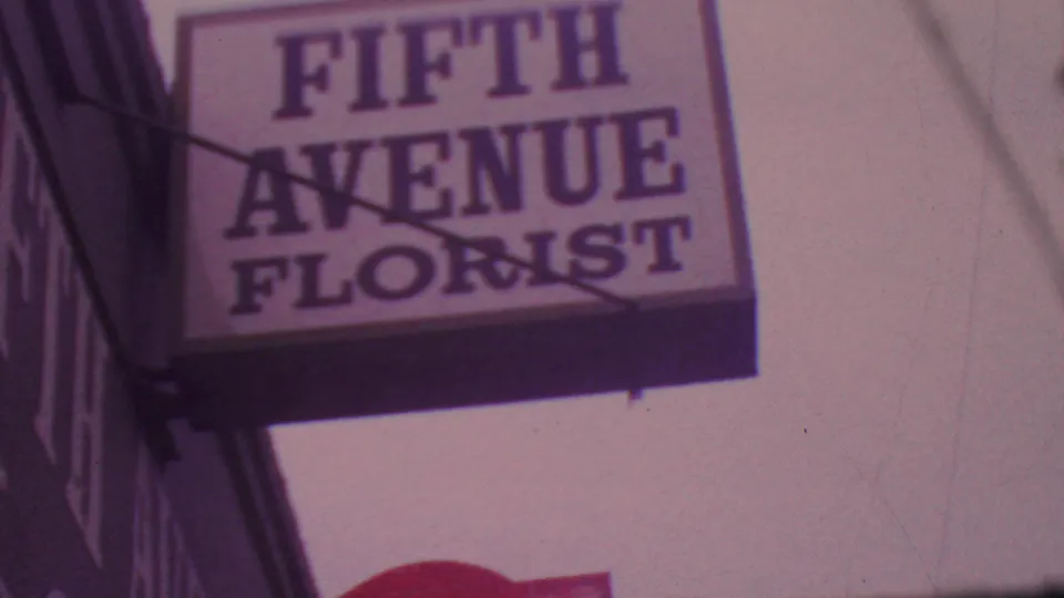 Close 8mm view of a Fifth Avenue Florist sign mounted above a sidewalk, with the building edge and tinted sky visible.