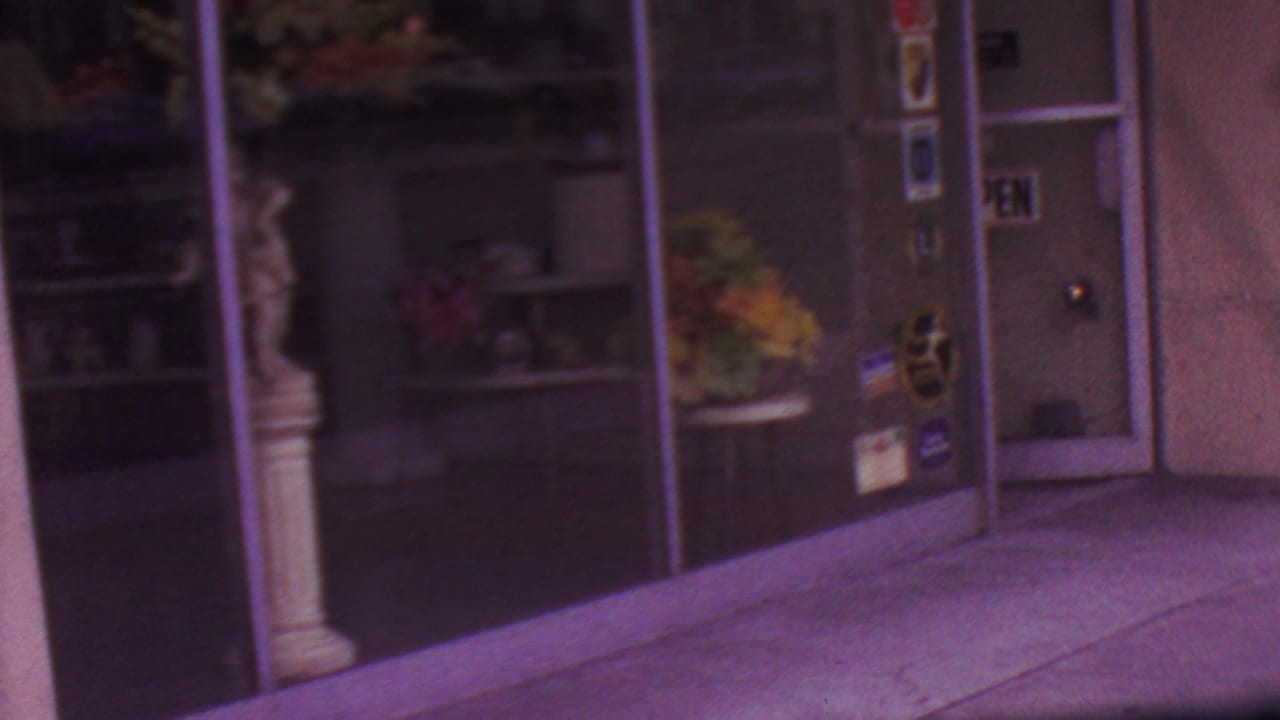 1965 Los Angeles home-movie frame showing reflective glass storefront doors, sidewalk pavement, and a classical-style statue beside the entrance.